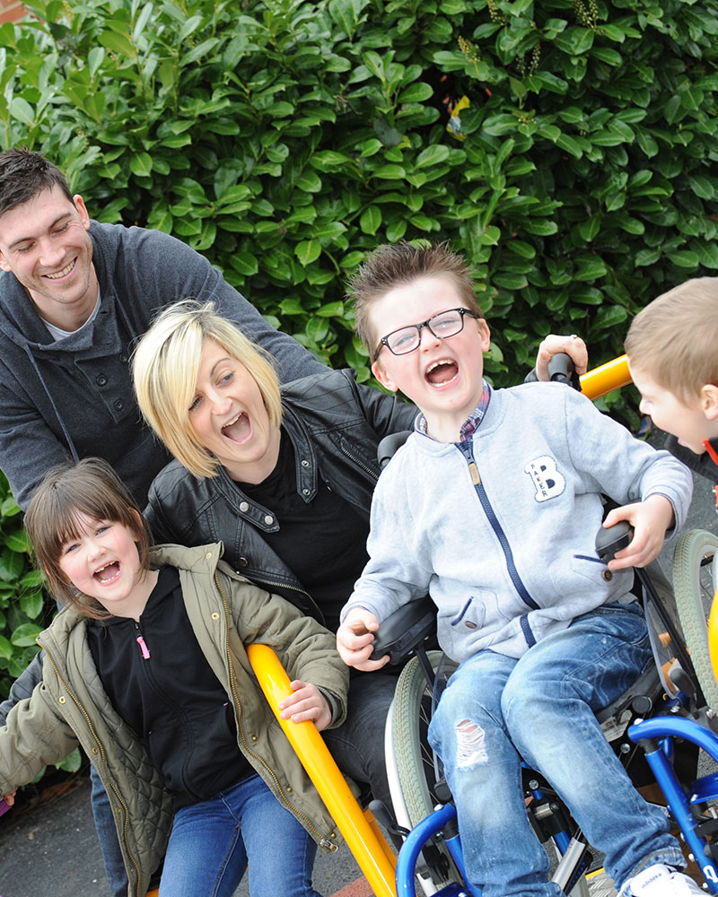 Family group are playing on an inclusive wheelchair accessible roundabout, they are all looking at the camera and smiling.